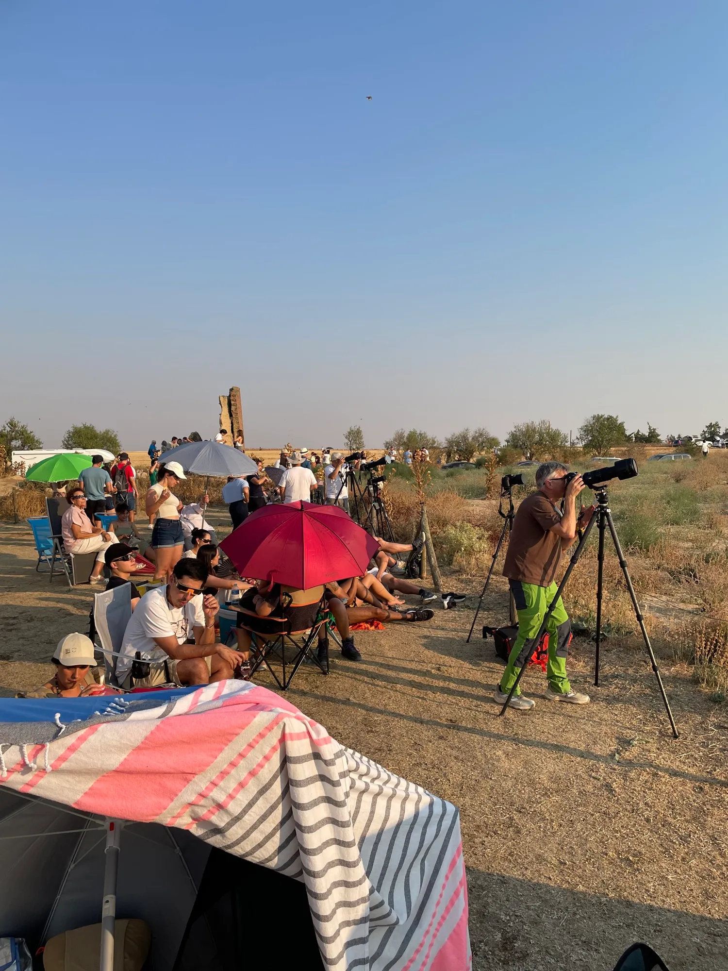 Gente con sombrillas y sillas esperando el eclipse, y un fotógrafo con teleobjetivo sobre trípode